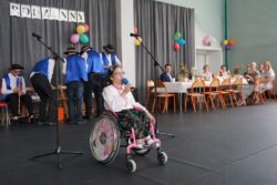 Dziewczynka na wózku inwalidzkim z mikrofonem w ręku pośrodku dużej sali. Za nią w tle siedzące przy stołach i stojące inne osoby.