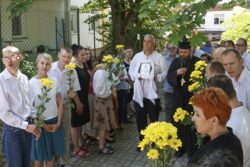 Duża grupa osób z kwiatami stojąca po obu stronach chodnika. Pomiędzy nimi duchowny i mężczyzna trzymający ikonę.