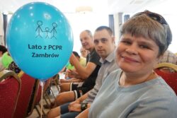 Siedząca na krześle kobieta trzymająca w rękach balonik z napisem: lato z PCPR Zambrów. W tle inne osoby. 