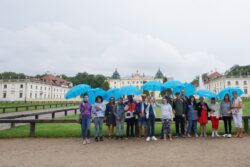 Grupa osób z parasolkami w ręku stojąca przed barokowym pałacem.