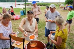 Kobiety nalewające zupę z dużego naczynia do miseczek. W tle jezioro i inne osoby.