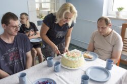 Kobieta krojąca tort stojący na stole. Przy stole dwóch siedzących mężczyzn.