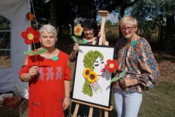 Trzy kobiety z kolorowymi kwiatkami w dłoniach stojące przy sztaludze na której stoi obraz.