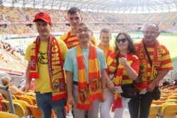 Grupa osób w pomarańczowo-żółtych koszulkach i szalikach stojąca na trybunie stadionu.