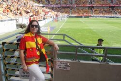 Kobieta w pomarańczowo-żółtych koszulce i szaliku stojąca na trybunie stadionu.