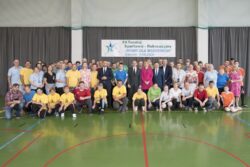 Zdjęcie grupowe bardzo dużej grupy ludzi ustawionych na dużej sali na tle szarej tkaniny i wiszącego na niej napisu XII Turniej Sportowo-Integracyjny.