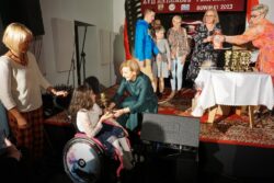 Kobieta wręczająca statuetkę dziewczynce na wózku inwalidzkim. Za nimi w tle grupa osób stojąca na scenie.
