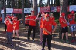 Grupa osób w czerwonych koszulkach i czapkach wykonująca wspólnie gimnastykę w lesie.