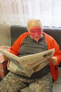Siedząca w fotelu kobieta z maseczką na twarzy czytająca gazetę.
