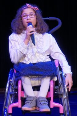 Dziewczynka na wózku inwalidzkim z mikrofonem w ręku.