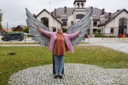 Kobieta stojąca przed makietą anielskich skrzydeł. W tle widoczny duży budynek.