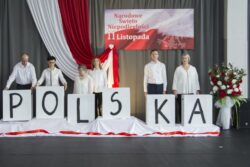 Grupa osób stojąca na tle biało czerwonej dekoracji z napisem Narodowe Święto Niepodległości 11 Listopada. Przed osobami napis Polska. 