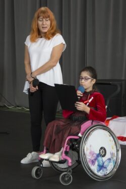 Dziewczynka na wózku inwalidzkim z mikrofonem w ręku i stojąca obok niej kobieta.