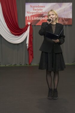 Dziewczyna z mikrofonem i teczką w rękach stojąca na dużej sali na tle biało czerwonych dekoracji.