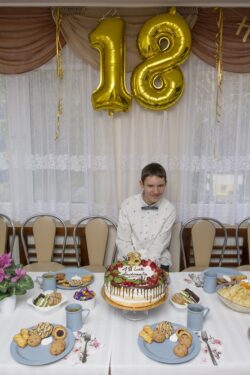 Siedzący za stołem chłopiec przed którym na stole stoi tort i talerzyki ze słodyczami. Nad chłopcem wiszą baloniki w kształcie liczby 18.