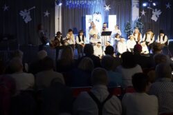 Grupa przebranych świątecznie osób z mikrofonami w dłoniach stojąca i siedząca na dużej sali na tle świątecznych dekoracji. Przed grupą siedzące osoby.