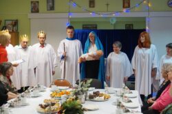 Grupa osób w białych szatach i innych scenicznych atrybutach stojąca naprzeciw świątecznie udekorowanego stołu przy którym siedzą inne osoby.