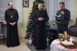 Dwóch duchownych i stojący obok nich przy udekorowanym świątecznie stoliku mężczyzna z mikrofonem w jednym a książką drugim ręku.