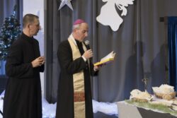 Dwóch duchownych stojący naprzeciw stołu na którym znajdują się opłatki. Jeden z duchownych trzyma w rękach mikrofon i książkę.