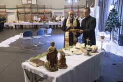Dwóch duchownych stojący naprzeciw stołu na którym znajdują się opłatki i inne przedmioty. Jeden z duchownych trzyma w rękach mikrofon i książkę.