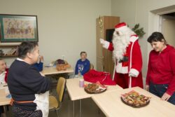 Mikołaj wchodzący do pomieszczenia w którym przy stolikach siedzą kobiety a na stolikach stoją ciastka.