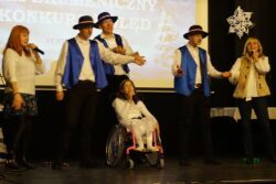 Dwie kobiety, trzech mężczyzn w kapeluszach na głowach i dziewczynka na wózku inwalidzkim stojący na scenie. 