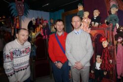 Trzech mężczyzn stojących w pomieszczeniu z różnymi lalkami teatralnymi.