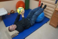 Mężczyzna wykonujący ćwiczenia z piłką na materacu w pozycji leżącej na plecach.