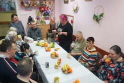 Grupa osób siedząca przy stole na którym leżą różne owoce i stoi sokowirówka przy której stoi kobieta.